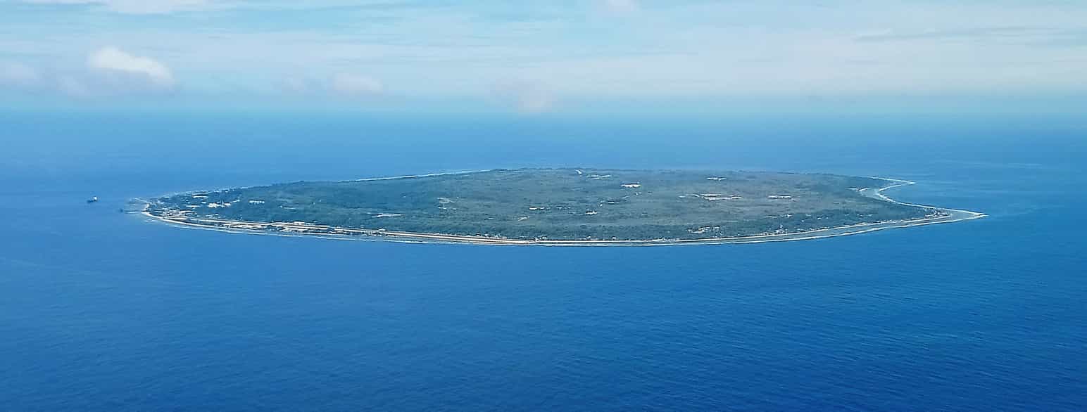 Luftfoto av en flat, grønn øy med en smal stripe sandstrand rundt. Blått hav. og himmel med noen få skyer.