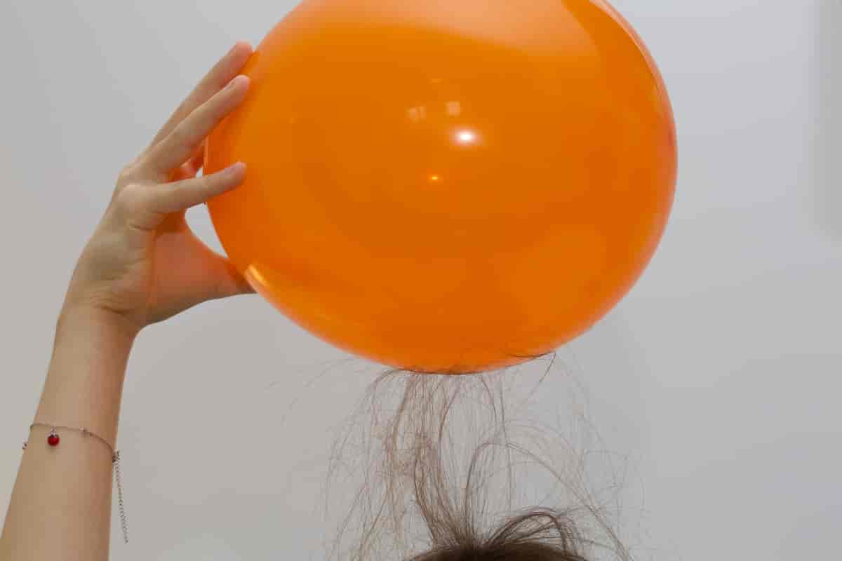 En hånd holder en oransje ballong over et hode. Håret på hodet har reist seg og festet seg mot ballongen.