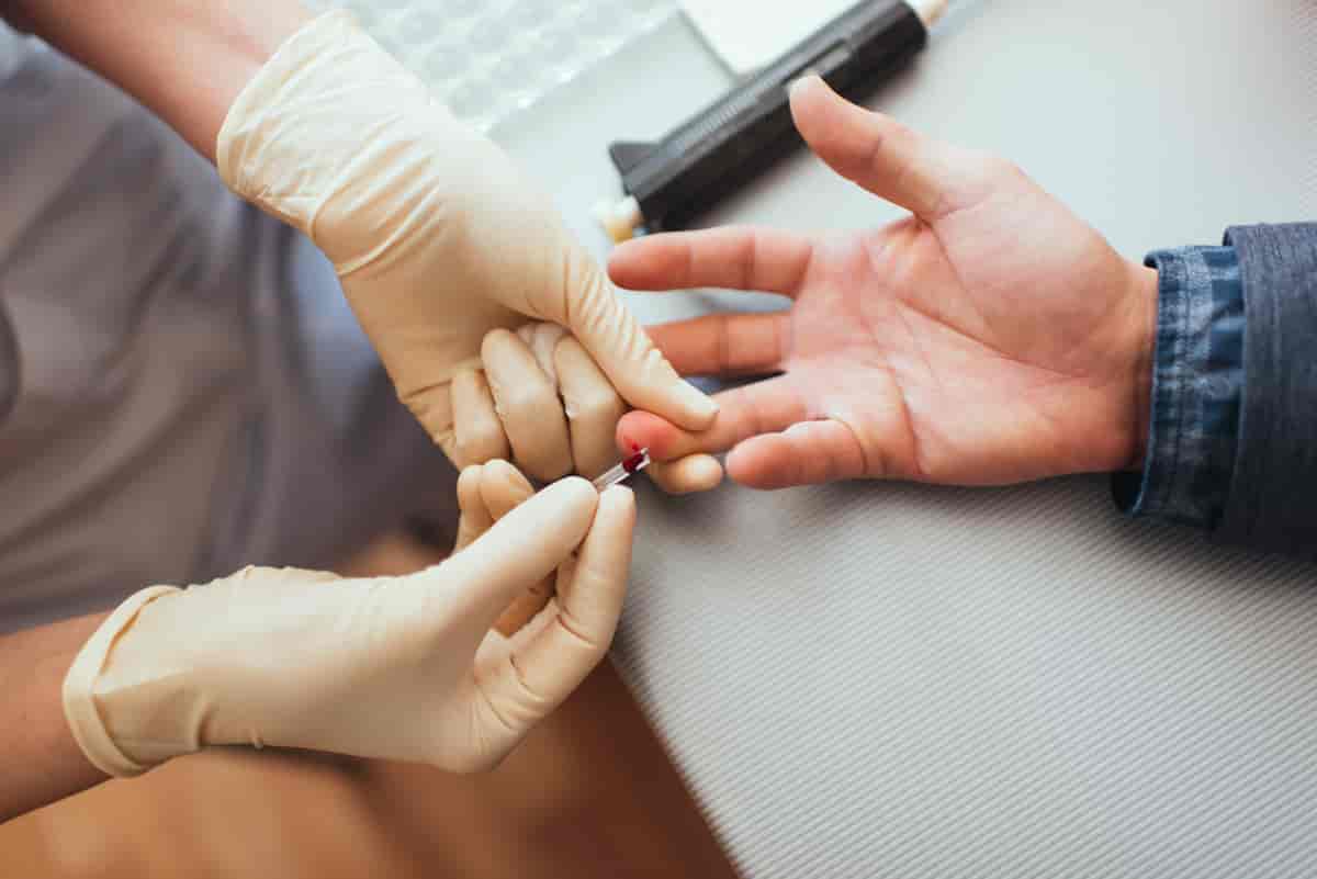 En person med gummihansker på hendene tar en dråpe blod fra fingeren til en pasient. 