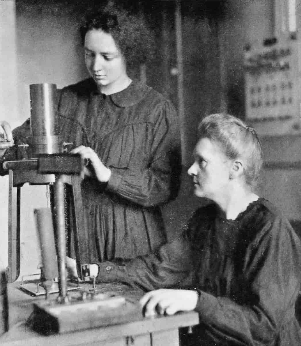 To kvinner ved et stort mikroskop. Irene Curie står ved mikroskopet. Marie Curie sitter ved et bord ved siden av og ser opp på Irene.
