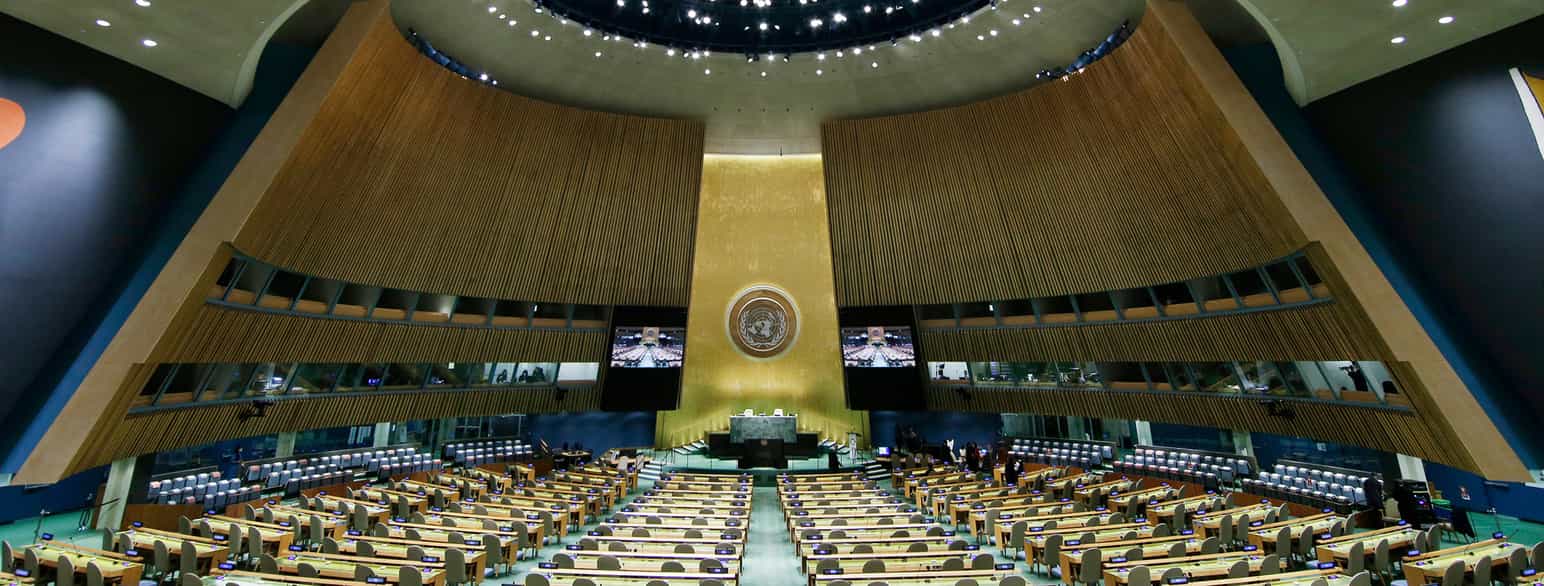 En kjempestor sal med rekker og rader av sitteplasser med bord foran. Forrest i salen er et podium med en stor skjerm på hver side. Midt mellom skjermene er FNs logo. Foto
