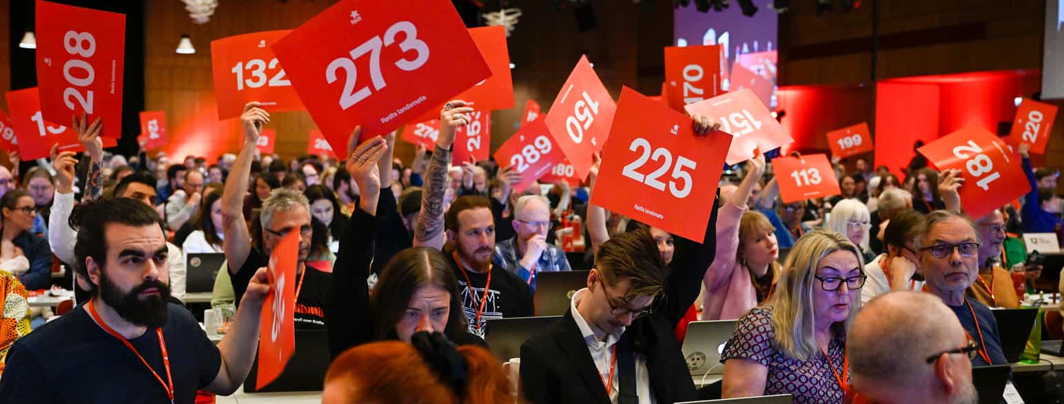 Foto fra en møtesal med mange mennesker. Flere av dem holder store, røde ark med hvite nummer på opp i lufta.