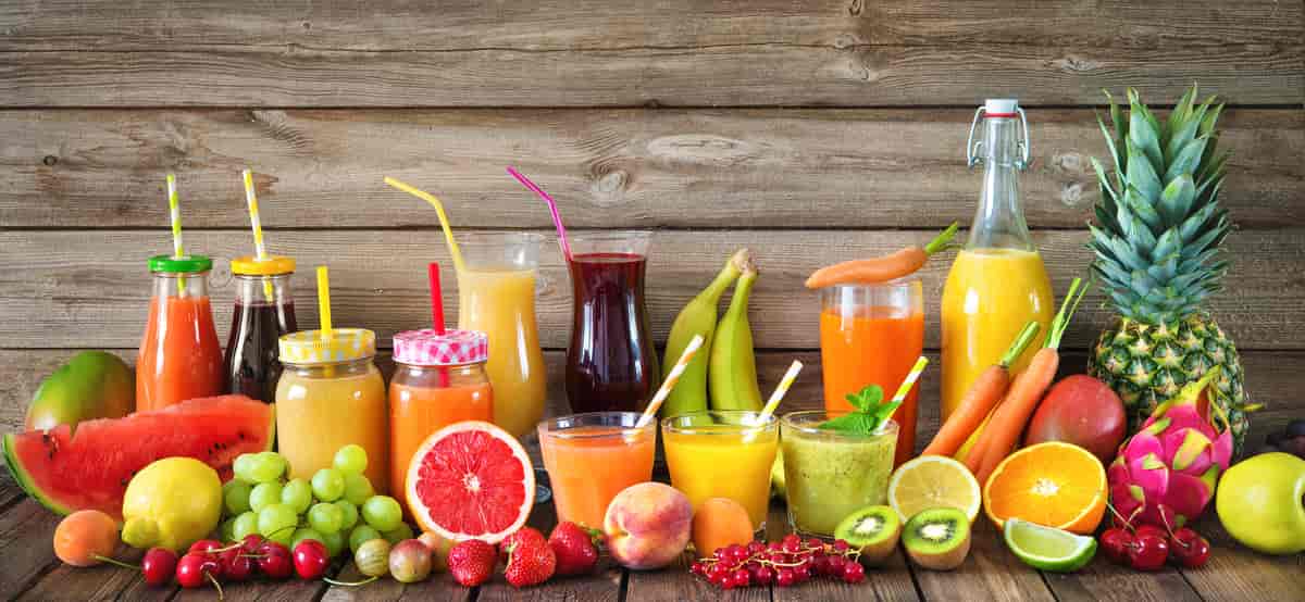 Diverse frukt og grønnsaksjuice i ulike glassbeholdere. Foran ligger forskjellige frukter, blant annet melon, drue, blodappelsin, jordbær, kiwi, morell og ananas. Foto