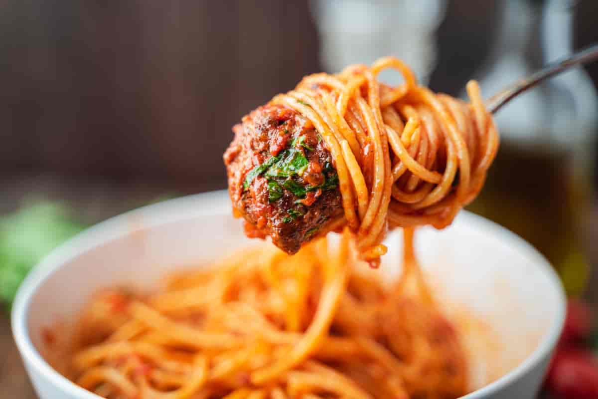 En hvit bolle med spaghetti med tomatsaus. Over bollen holder holder noen en gaffel med spaghetti snurret rundt, og saus og kjøttbiter i enden. Foto