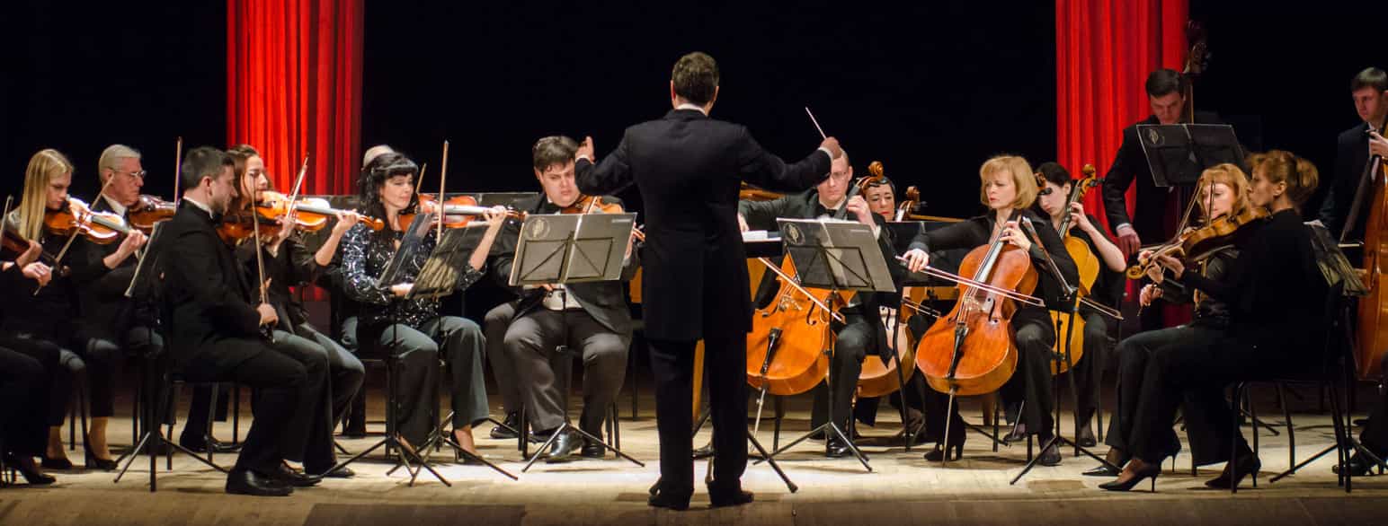 På bildet er en dirigent med ryggen til et lite orkester. Man ser musikere som spiller fiolin, cello og bratsj. Dirigenten holder en taktstokk i høyre hånd. 