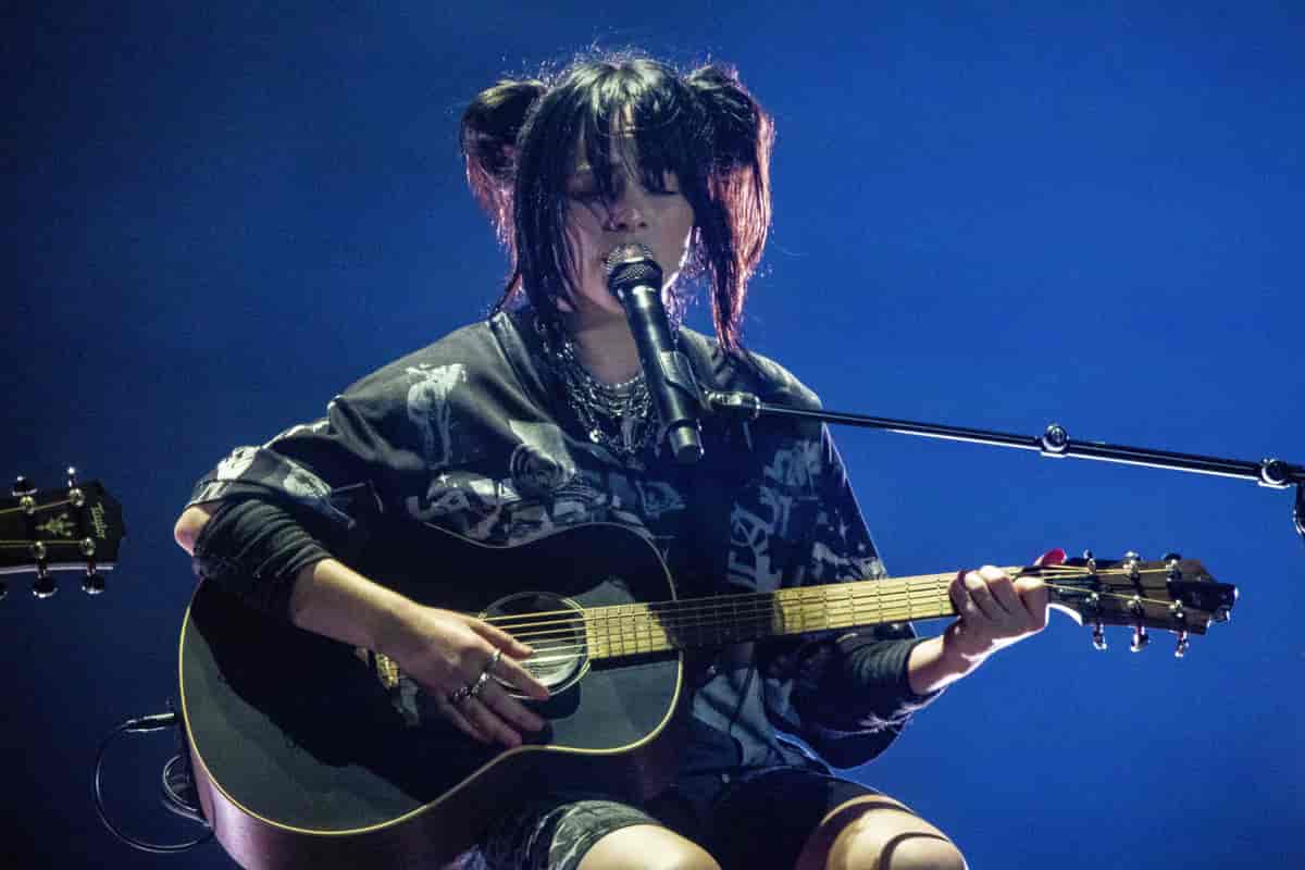 En ung kvinne sitter på en stol. Hun spiller gitar og synger inn i en mikrofon.