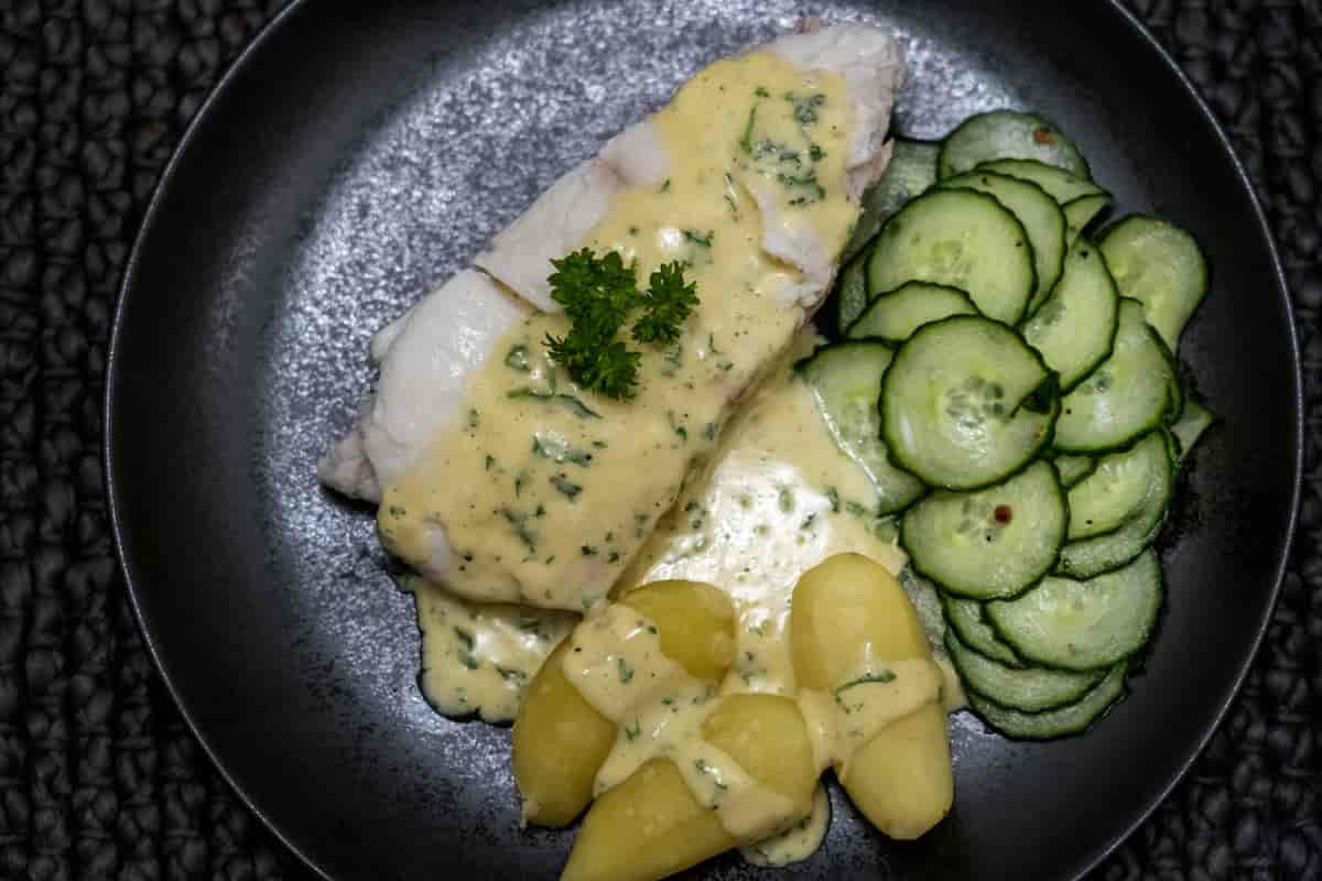 Et stykke hvit fisk på en mørk tallerken. Ved siden av er det skivet agurk og tre små kokte poteter. Over fisken og smøret er en lys gul saus med biter av hakket persille. Foto