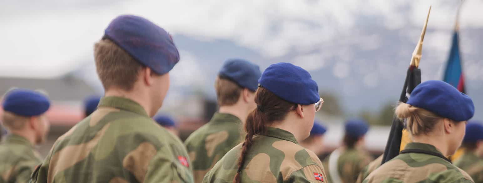 Foto av fem soldater i uniformsskjorter i grønn-brun kamuflasje-farge står oppstilt med ryggen til kamera. Alle har mørkeblå bereter. Tre er kortklipte, to har langt hår som er satt opp i hestehale eller flette.