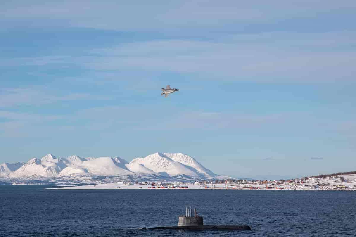 Et vinterlandskap med fjord og høye, bratte fjell dekket av snø. I lufta er det et militærfly. En ubåt ligger i overflaten av sjøen.