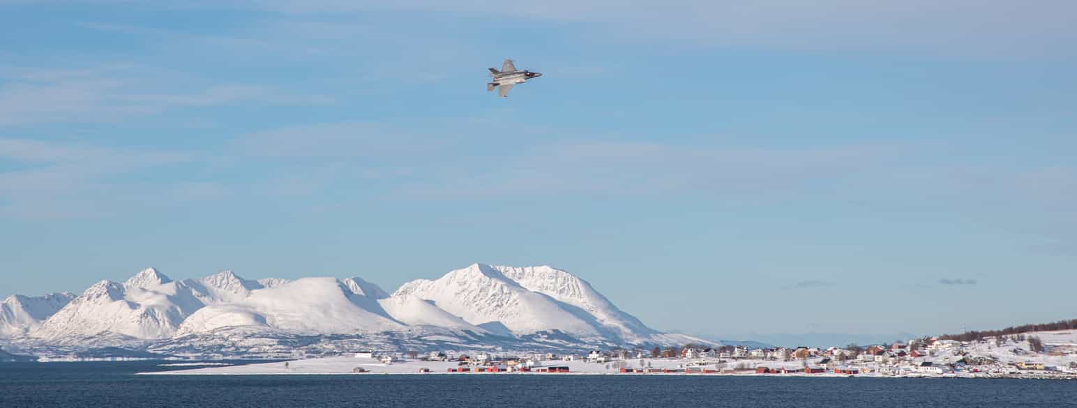 Vinterlandskap med fjord og høye, bratte fjell. Fjellene er dekket av snø. I lufta er det et militærfly. Foto