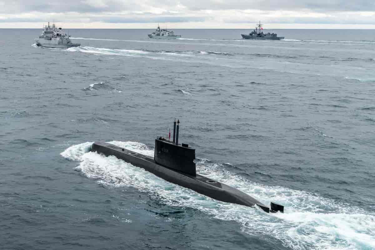 En ubåt i fart på overflaten av sjøen så vannet skummer rundt den. I horisonten i bakgrunnen er det tre  andre militære båter.