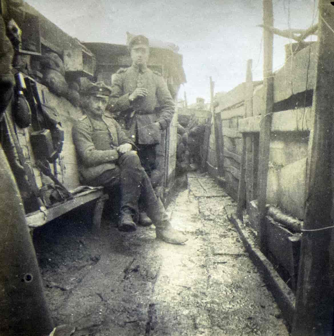 Svarthvitt foto av to menn i uniform i en konstruksjon av treplanker. Til høyre i bildet er en gjerdelignende vegg. Til venstre en vegg med en smal benk hvor det henger forskjellig utstyr. Den ene mannen sitter på benken, den andre står ved siden av. 
