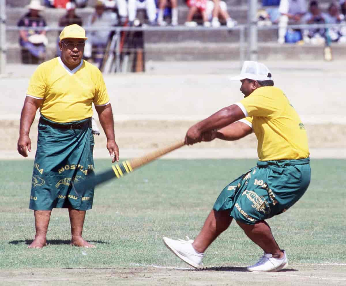 To menn i spillerdrakter som består av gul t-skjorte og grønn sarong. Den ene ser på at den andre svinger et balltre. I bakgrunnen sees en tribune med noen få tilskuere.