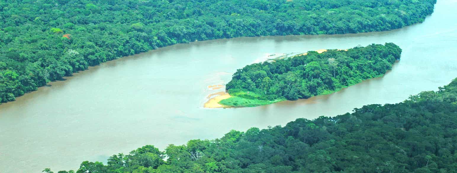 Flyfoto. En elv med stille vann og en grønn øy i midten renner gjennom en tett grønn skog. Foto