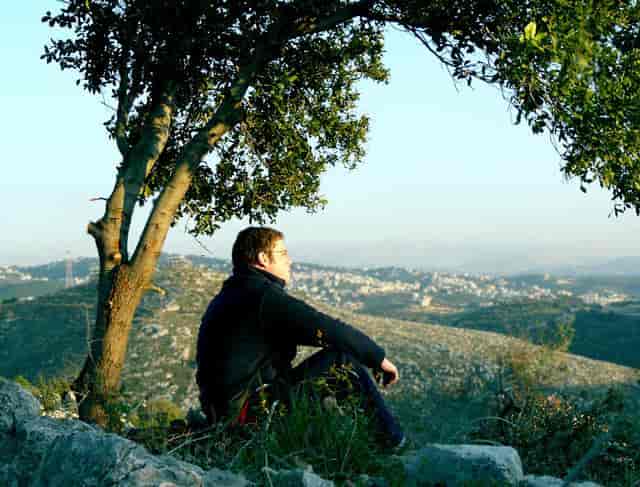 En mann sitter utnder et tre og ser utover et fjellandskap.