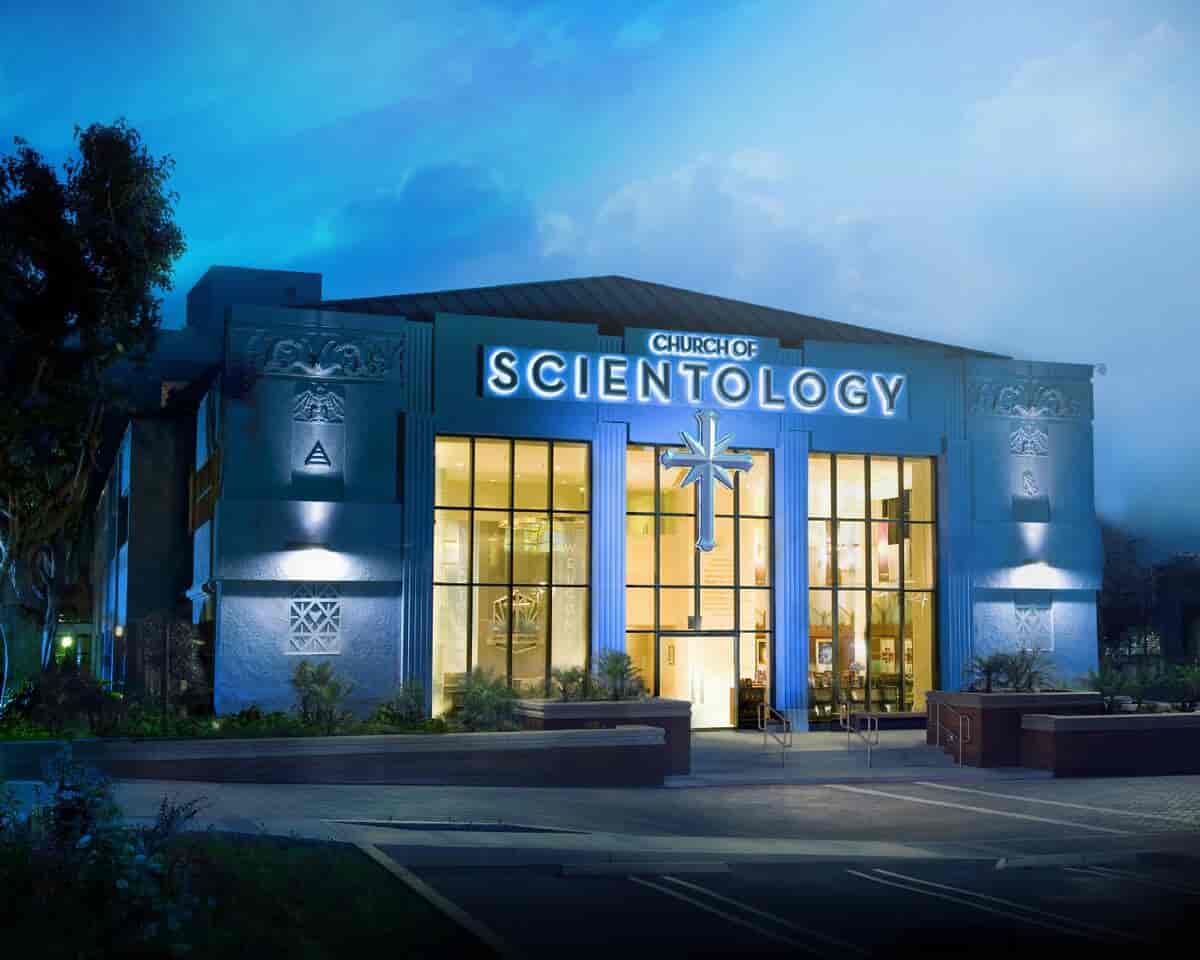 En stor, hvit bygning med et kors over inngangsdøren. På veggen står det Church of Scientology med store bokstaver. Nesten hele veggen foran er vinduer. Ute er det kveld eller natt, det lyser inne i bygningen.
