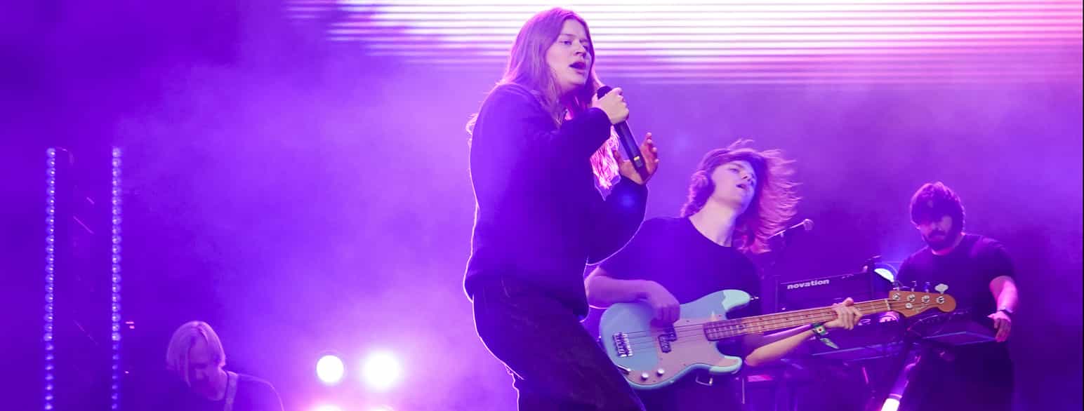 En jente med langt hår som synger inn i en mikrofon på en scene. Til høyre for henne ser man en mannlig gitarist og en som spiller keyboard.