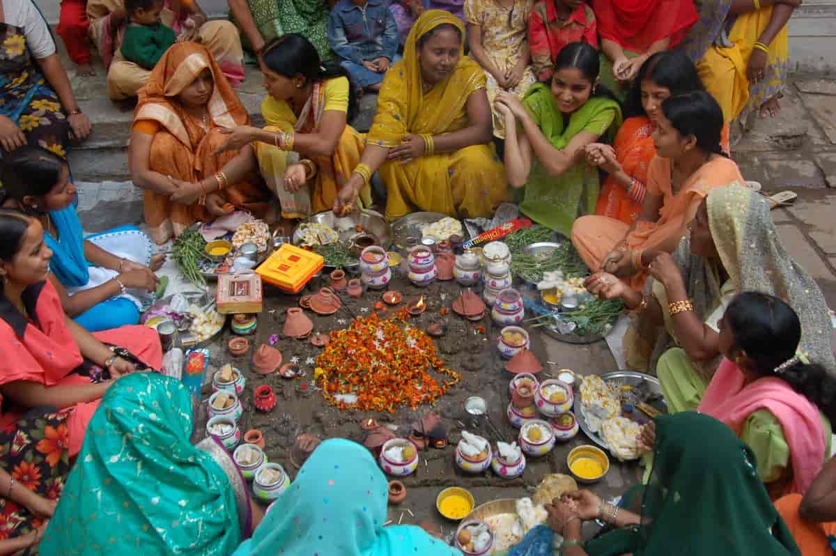 Foto av kvinner i fargerike klær som sitter på bakken i en ring. Midt i ringen er det små skåler og kopper og tallerkner med mat. Noen av kvinnene har sjal.