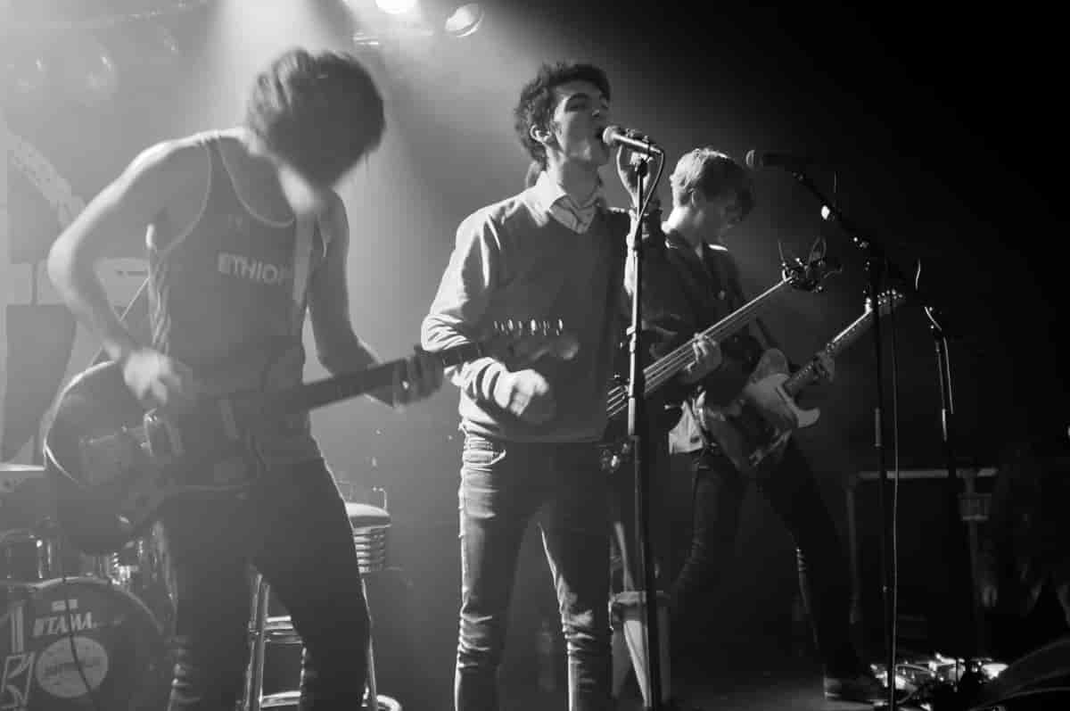 Tre unge musikere på en scene. Bildet er i svart/hvitt. To har gitar og en sanger i midten synger inn i en mikrofon.