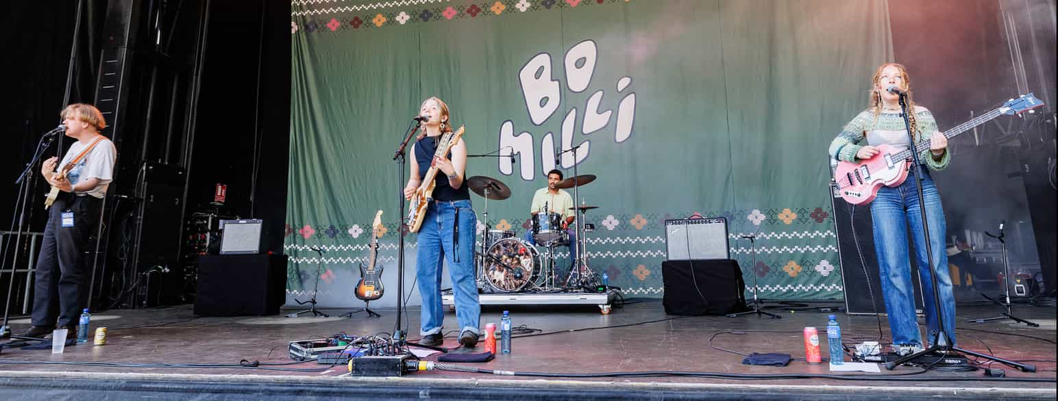 En ung musiker som spiller giar og synger. Hun står på en scene. Bak på veggen står det Bo Milli med store bokstaver. På høyre side er en annen kvinnelig musiker som spiller gitar, og på venstre side er det en mannlig musiker som spiller gitar.