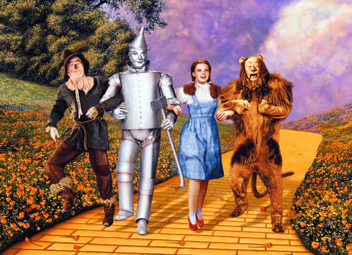 En jente med fletter og blå kjole står i midten. Til venstre står en mann kledt ut som en tinnmann, bak til høyre står en mann kledd ut som et fugleskremsel og til høyre står en mann kledd ut som en løve.