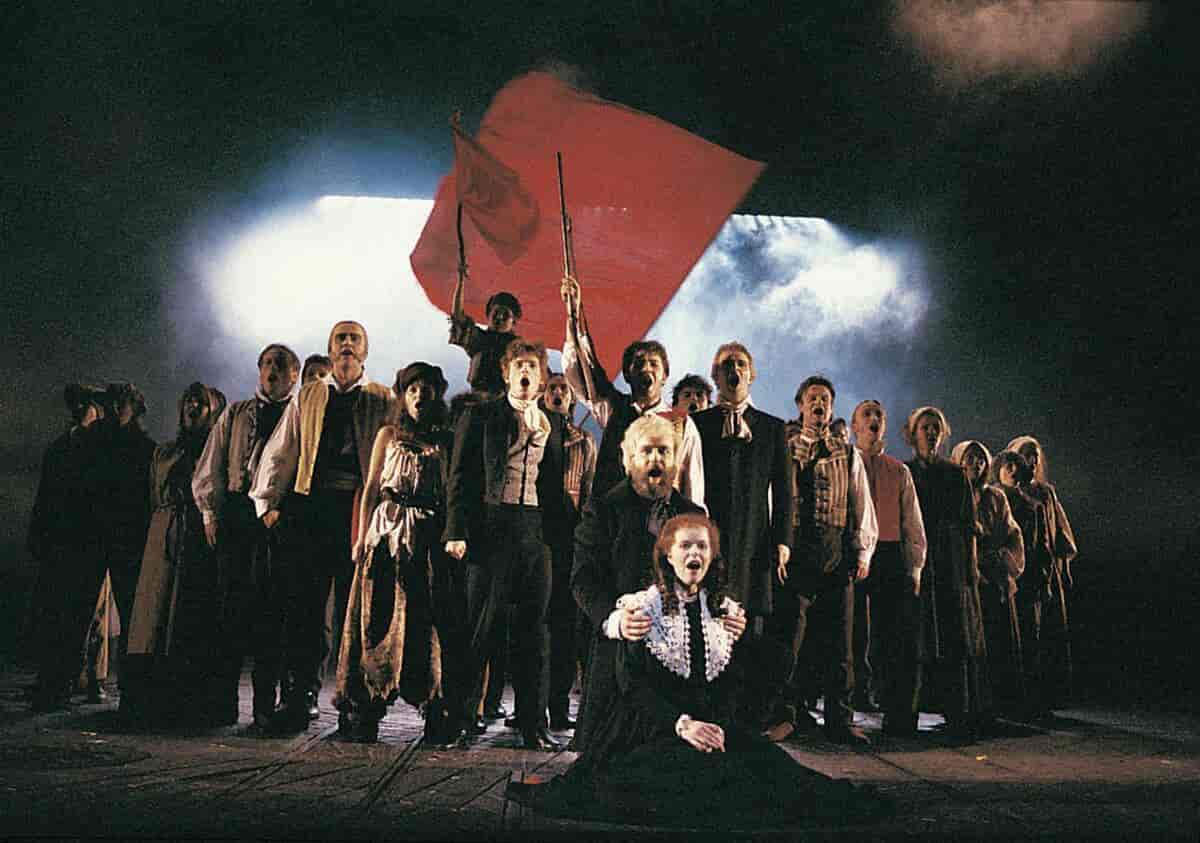 Skuespillere på scenen som synger. Et stort, rødt flagg i bakgrunnen. Foran sitter en mann på huk og holder rundt en ung kvinne som sitter.