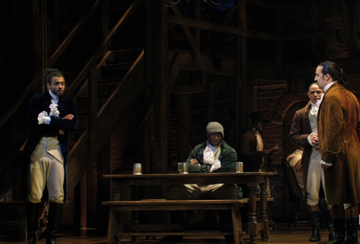 Fire menn på en scene. En av dem sitter ved et bord, de andre står rundt. Alle er kledt i historiske klær fra 1700-tallet.