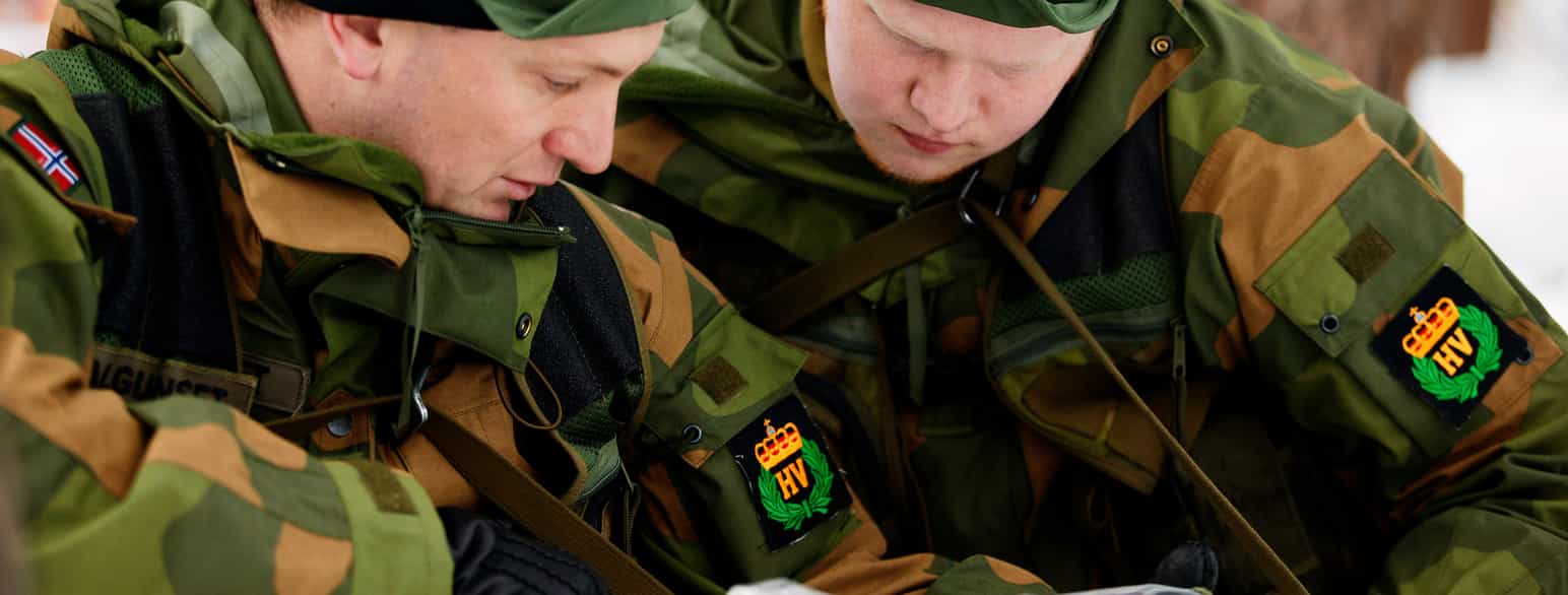 Foto. Nærbilete av to soldatar i grøne uniformar som ser ned på noko som den eine held. På erma har dei merke der det står HV.
