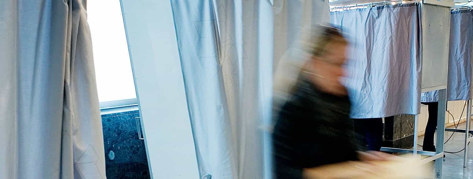 Foto av en person som beveger seg vekk fra et avlukke med grå gardiner foran. Personen er i ufokus på grunn av farten i bevegelsen. Ved siden av avlukket personen kommer ut fra er det et likt avlukke med gardinene trukket for. 