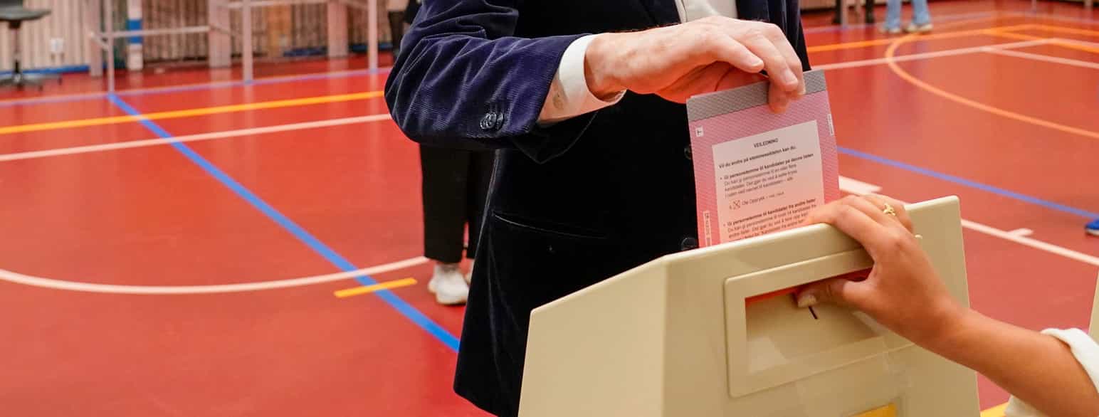 Foto. Nærbilde av armen og hånden til en person som legger en stemmeseddel i en valgurne. Hånden til en annen person holder på valgurnen.