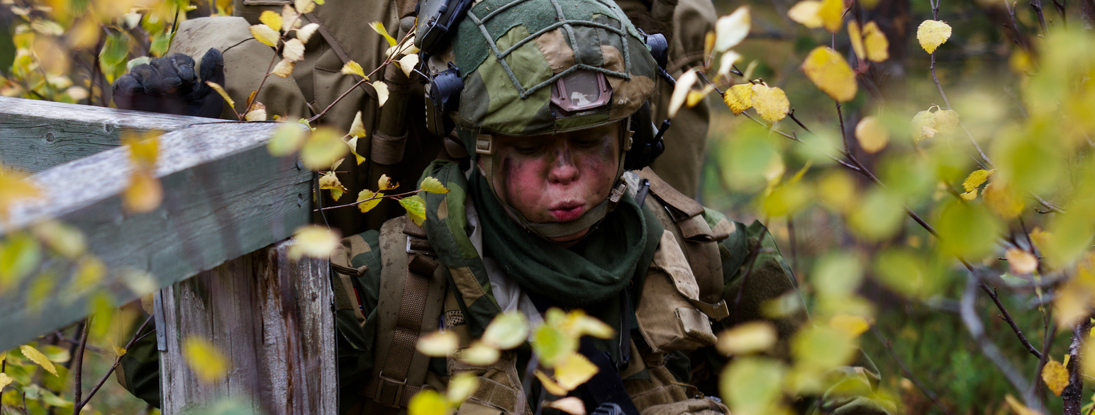 Foto av en soldat i grønn feltuniform med hjelm, stor ryggsekk og kamuflasjemaling i ansiktet holder i et plankegjerde mens hun er på vei gjennom et kratt med tynn løvskog.