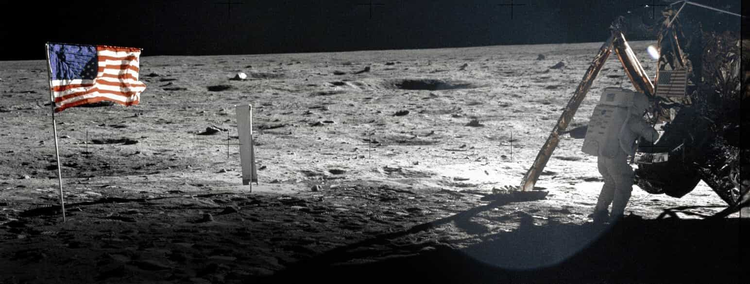 Overflaten på månen med verdensrommet i bakgrunnen. Til venstre et amerikansk flagg plantet i bakken. til høyre ser man en del av et romfartøy og en astronaut som er på vei inn i fartøyet.