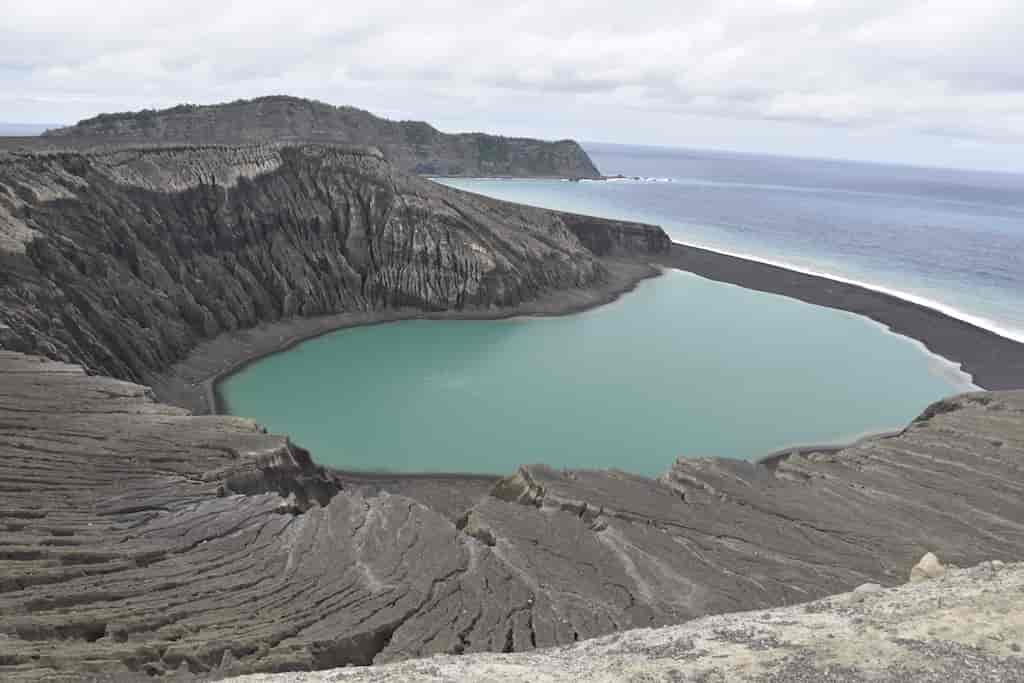 Utsikt over en flat fjelltopp med et krater av gråsvart lava. I krateret er det en dam av turkist vann. I bakgrunnen, hav og en øy med noen få trær.