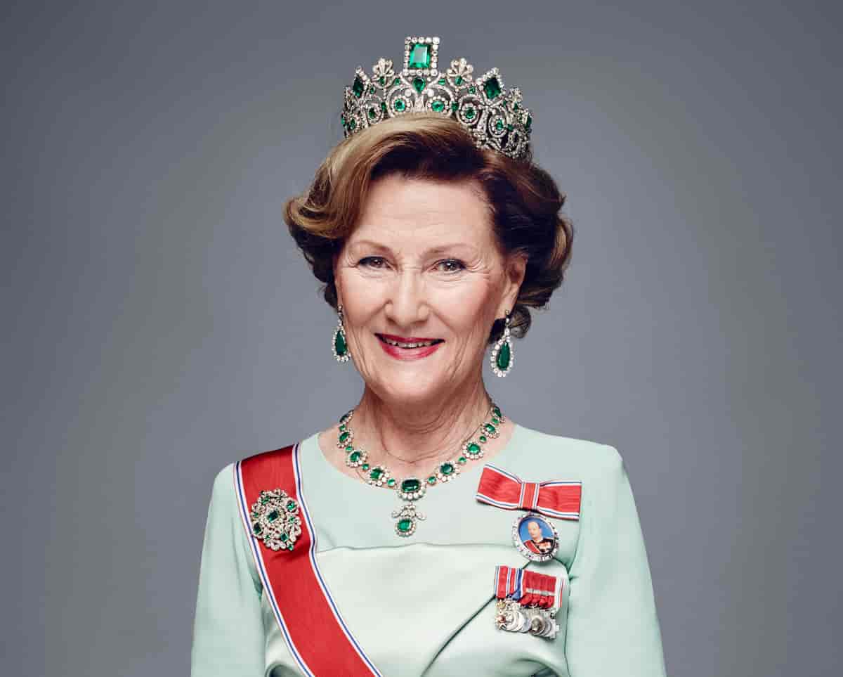 Dronning Sonja har en stor tiara med grønne steiner, rødt ordensbånd over en lys grønn kjole og en medaljong med bilde av kong Harald 5. 