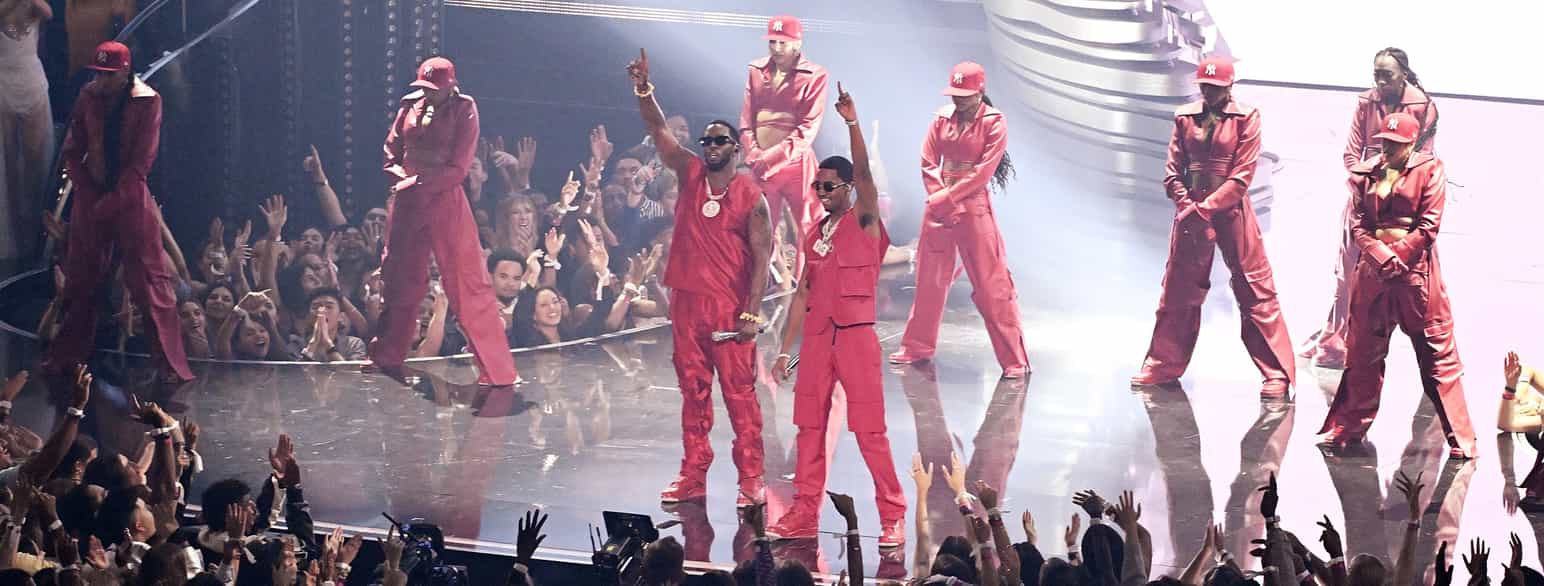 To mannlige rappere i midten på en scene som tar i mot applaus fra publikum. De er helt kledt i rødt. Rundt dem er det dansere. Alle danserne har caps.