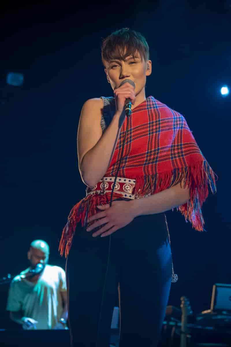 En kvinne med at samisk sjal over skulderen synger inn i en mikrofon. I bakgrunnen står en mann bak et keyboard. Han synger også inn i en mikrofon. Foto