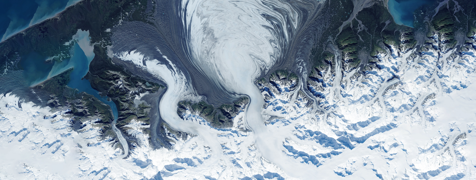 Luftfoto av en kyst tatt rett ovenfra. Landet har spisse fjelltopper, dekket av is og snø. Ytterst ved sjøen er det noen bare fjelltopper. Midt i bildet renner en iselv ut i havet. Foto
