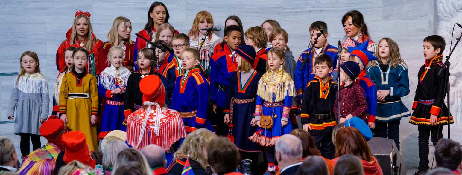 Et barnekor med barn kledd i forskjellige samiske kofter inne på Oslo rådhus. Dirigenten står med ryggen til og har på rød kyse og et rødt og hvitt sjal med frynser. I publikum sitter flere voksne, noen av dem har også kyser og sjal. Foto