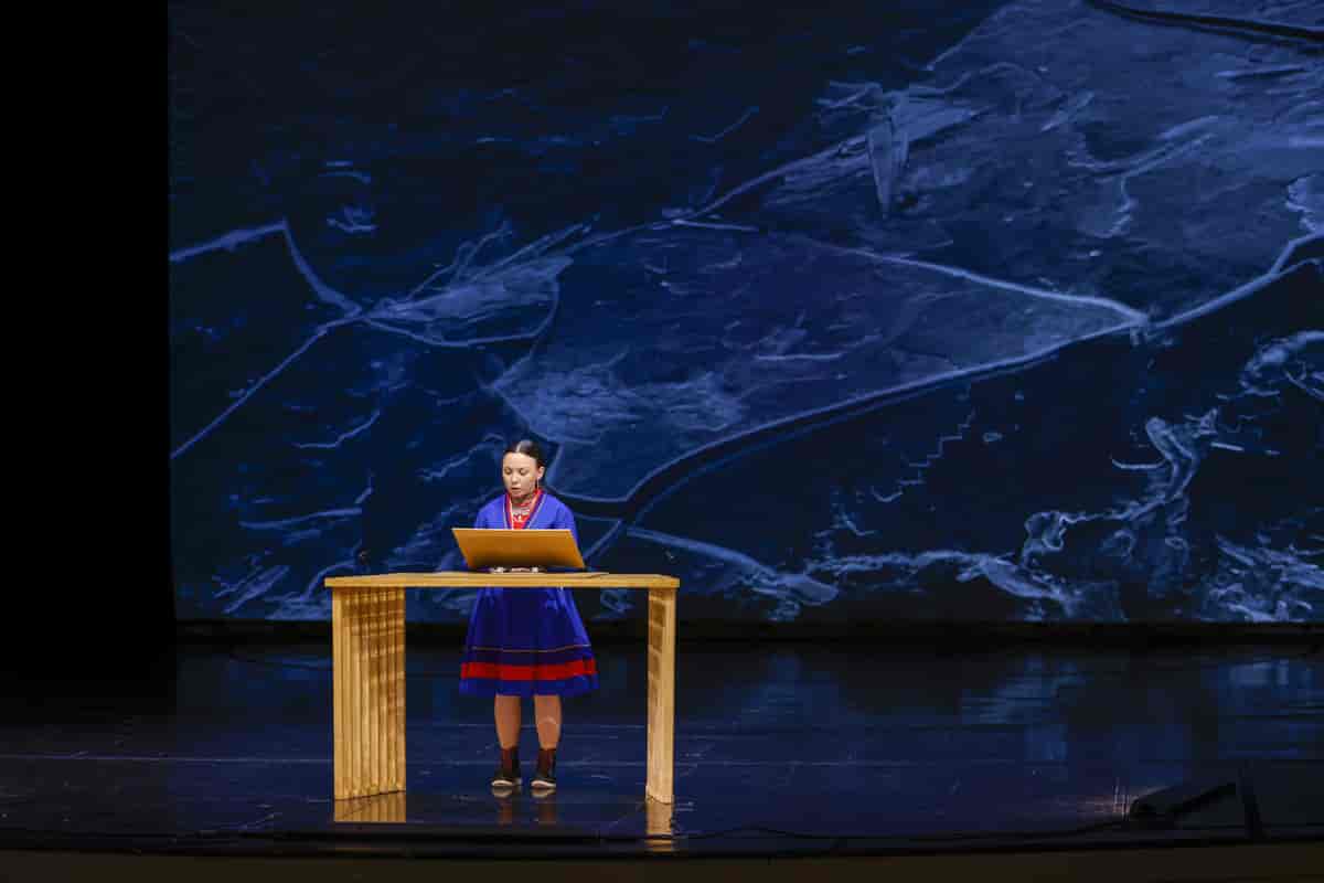En ung kvinne i samisk kofte står bak et bord på en stor scene og ser ned på en skjerm hun har foran seg. Gulvet er mørkt og blankt, og gjenspeiler det mørkeblå scenebakgrunnen. Foto