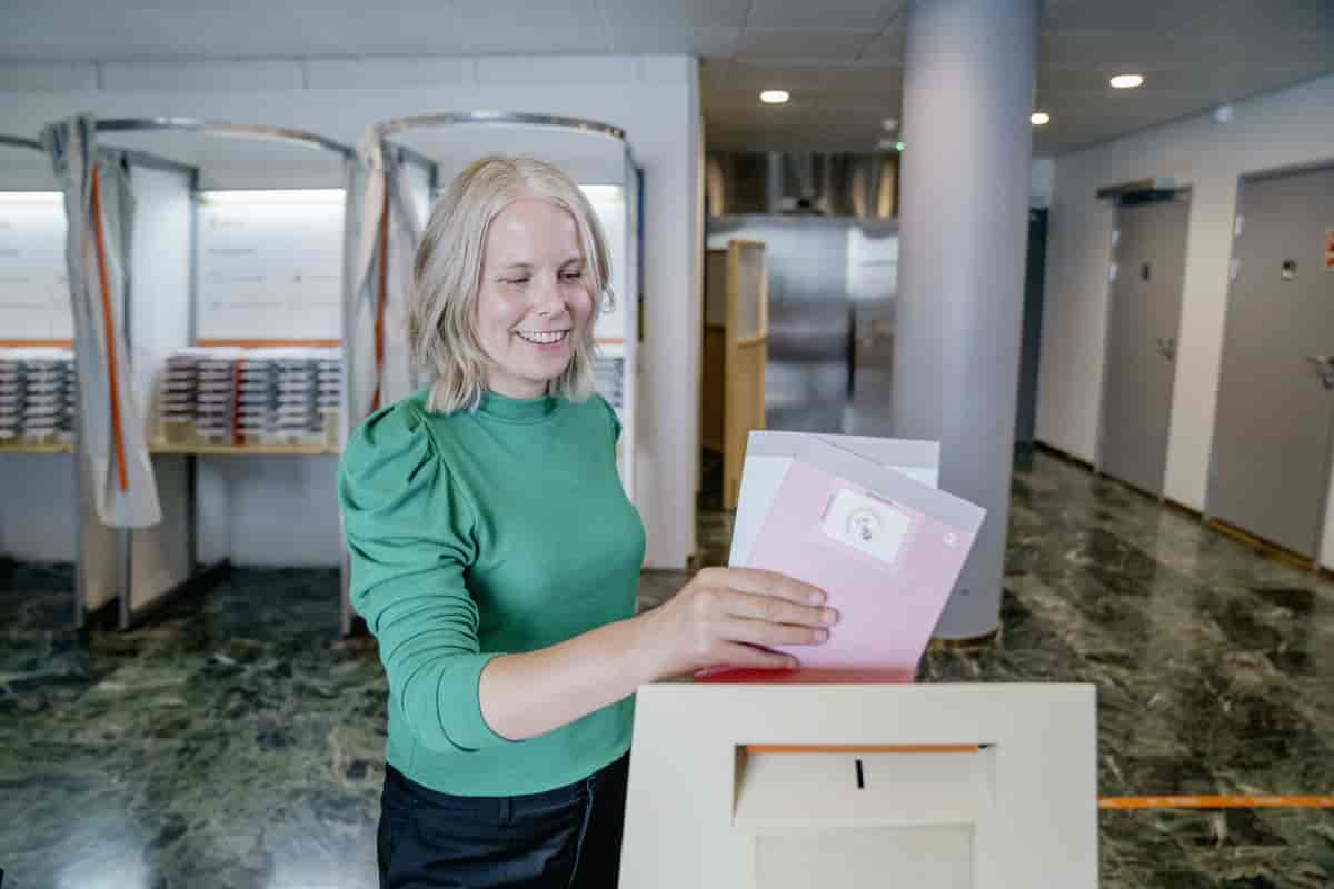 Foto av Bergstø som holder fram to stemmesedler som hun skal putte i en valgurne. I bakgrunnen er det båser med stemmesedler og gardiner til å trekke for når man skal velge.