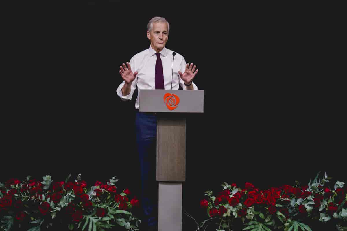 Foto av en mann med hvit skjorte og burgunder slips står bak en talerstol og snakker mens han holder begge håndflatene opp. Scenen er pyntet med røde roser på hver side av talerstolen. Bakgrunnen er helt mørk. Foto