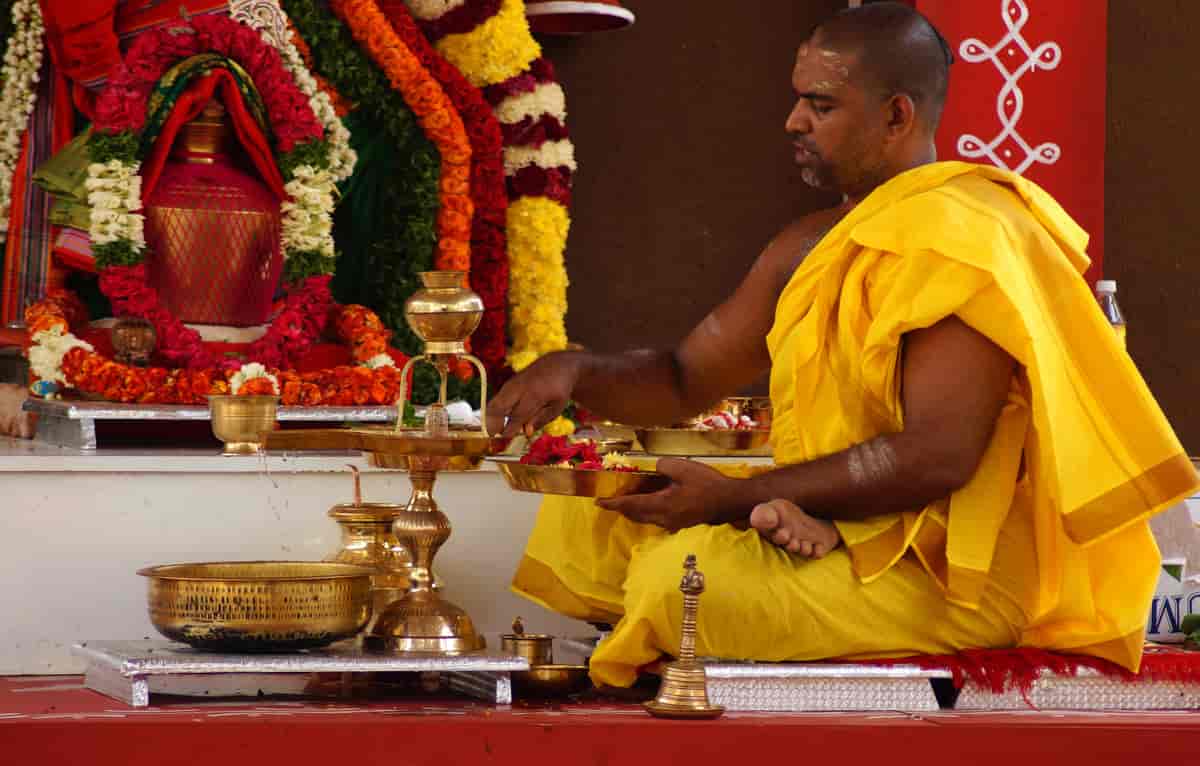 En indisk mann sitter med bena i kors foran et lavt bord med et fat i gull med blomster i mange farger. Det er også andre ting av gull, som en lysestake og en bolle. Han har på seg en tradisjonell gul drakt. Bak ham kan man se fargerike girlandere som henger på veggen