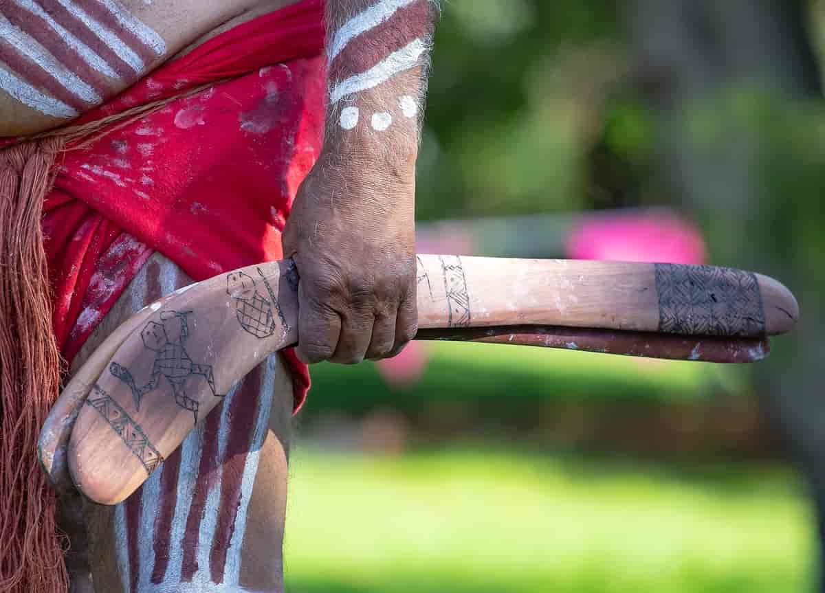 Foto av kroppen til mann som holder to bumeranger. Han er malt med striper og prikker i hvitt, og har bundet et rødt klede rundt livet.