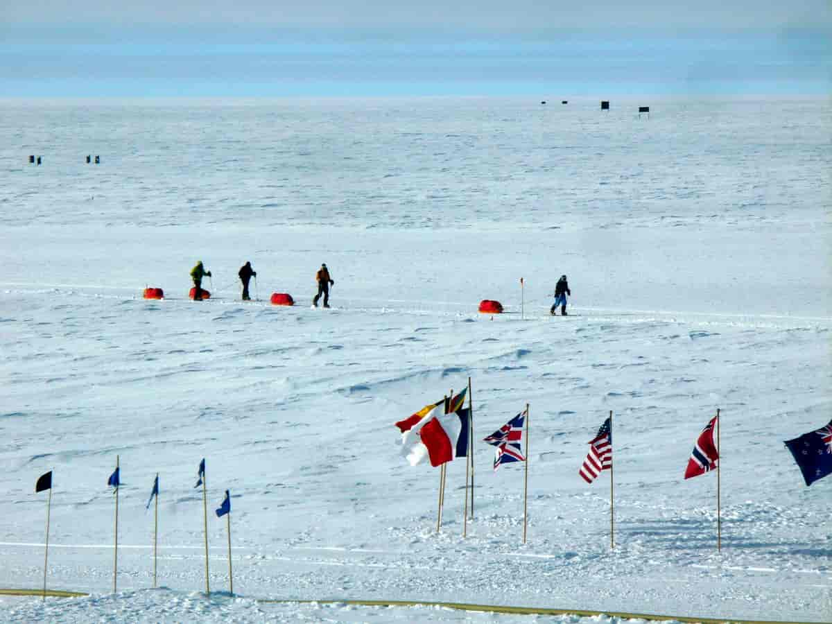 Bildet viser et flatt landskap som er dekket av snø. I forgrunnen er mange lands flagg plantet, blant annet det norske flagget. Midt i bildet går fire mennesker på ski med pulker på slep.