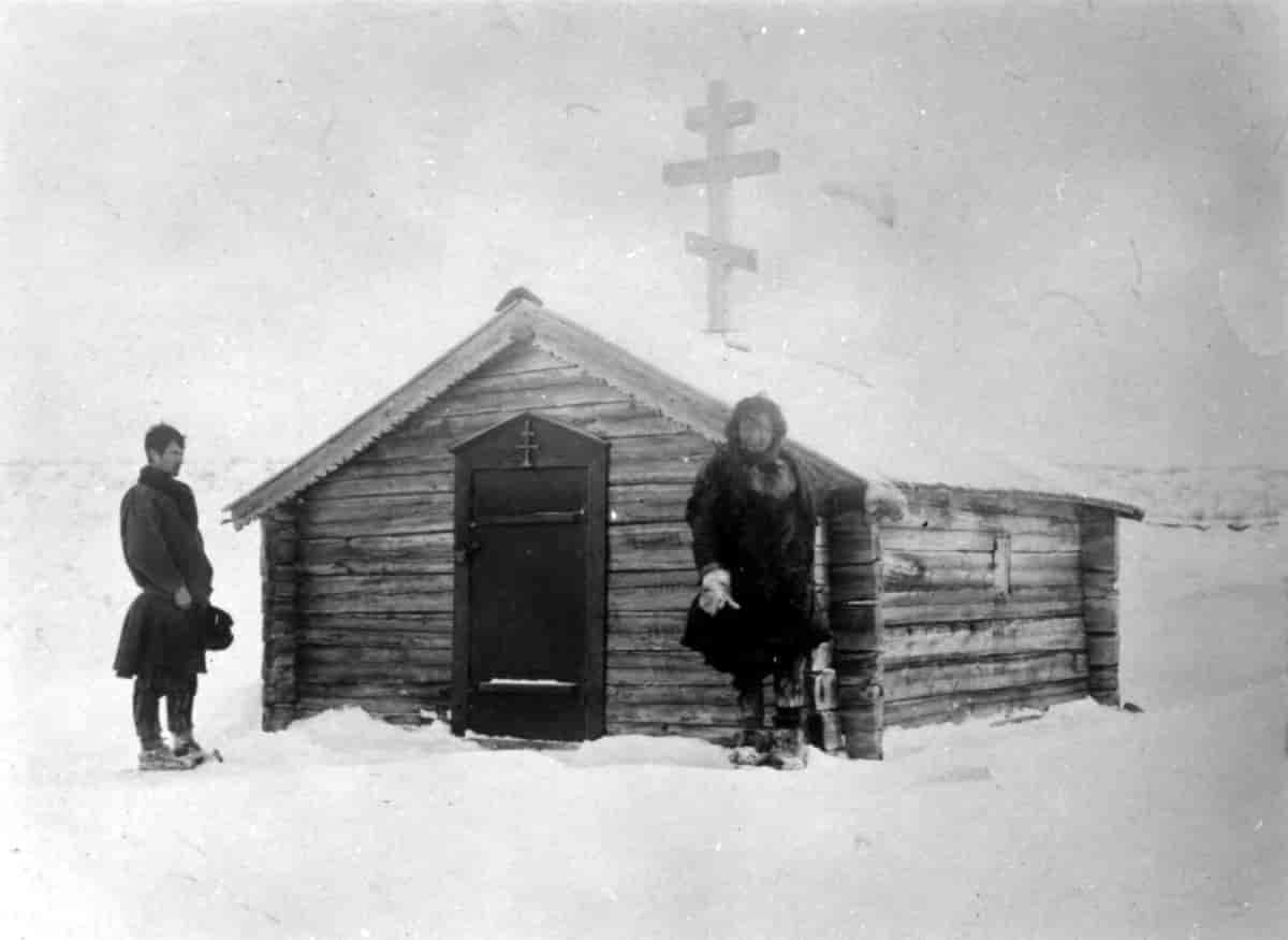 To personer utenfor en liten bygning av laftet tømmer. På taket er det et ortodoks kors med tre pinner på tvers. Svart-hvitt foto