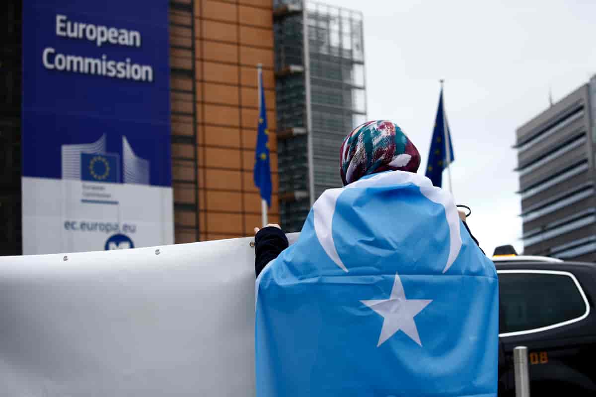 Foto av en person med et lyseblått flagg med hvit sigd og stjerne knytt rundt skuldrene og et skaut på hodet står med ryggen til kamera. På en bygning ved siden av står det European Commission.