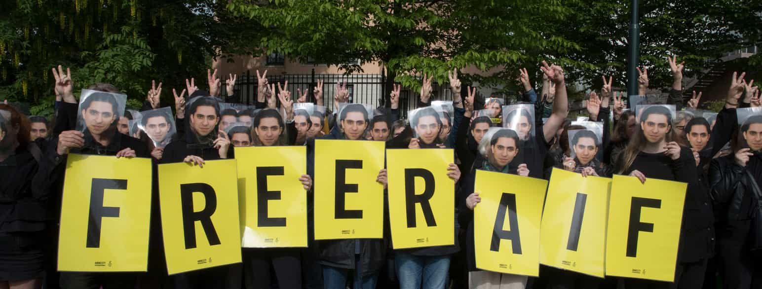 En gruppe mennesker holder gule plakater med én bokstav på hver, og alle holder et utklipp av ansiktet til en person foran sine egne ansikter. De som ikke holder plakater holder opp en hånd og viser fredstegnet med to fingre opp i lufta. Bokstavene på plakatene staver ordene «FREE RAIF». Foto