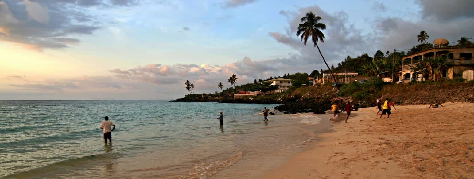 En strand i solnedgang. Noen personer spiller fotball i vannkanten. Langs kysten i bakgrunnen er det noen bygninger og noen få palmer. Foto