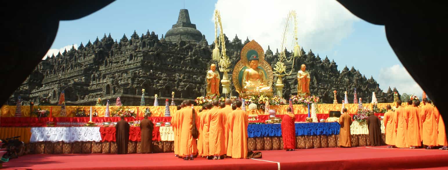 Mange buddhistiske munker i oransje kapper står med ryggen til.  Et stykke foran dem er det en veldig stor Buddhastatue av gull. Det er pyntet med blomster rundt og foran statuen. 