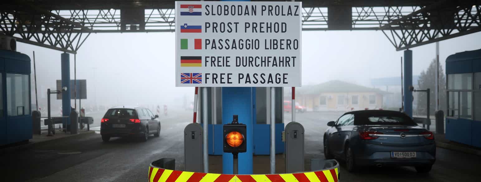 En grensestasjon med plass for biler å kjøre gjennom. På en stolpe mellom to filer henger et skilt hvor det står «FREE PASSAGE» på fem språk. Foran hvert språk er det flagg, fra øverst: Kroatia, Slovenia, Italia, Tyskland, Storbritannia. Foto